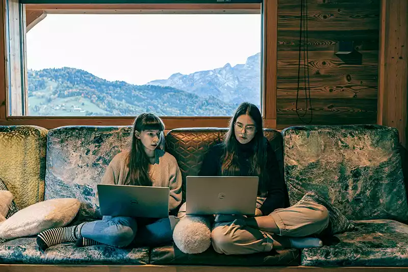 Zwei Frauen sitzen auf einem Sofa und schauen gemeinsam in ihre Laptops