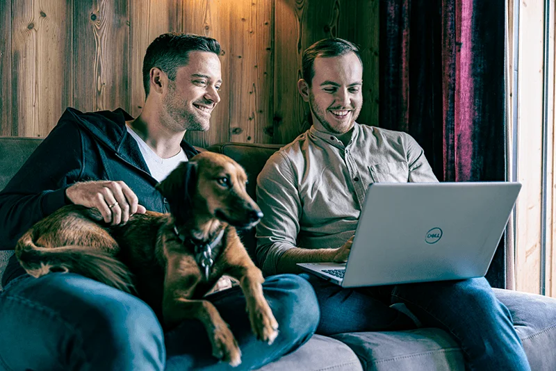 Männer sitzen mit einem Hund auf einem Sofa und schauen in einen Laptop