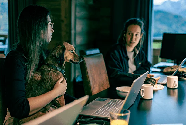 Zwei Frauen mit einem Hund auf dem Schoß sitzen an einem Tisch mit Laptops und Tassen